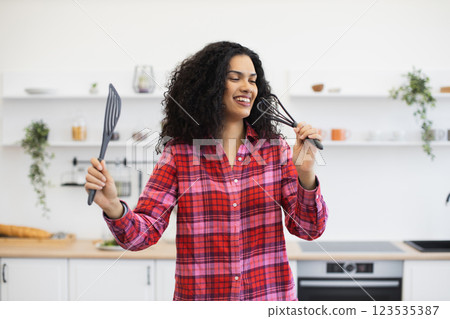 Young African woman, wearing vibrant plaid shirt, standing in kitchen, holding utensils, smiling energetically, expressing creativity and joy. Young African woman, wearing vibrant plaid shirt, standing in kitchen, holding utensils, smiling energetically, expressing creativity and joy. 123535387