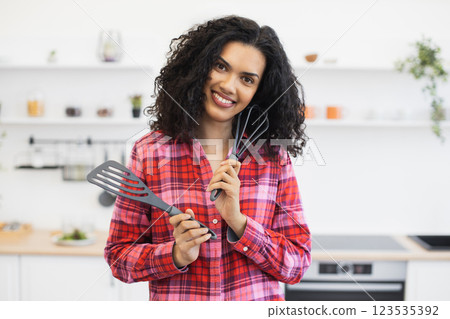 Young African woman with curly hair holding kitchen utensils in modern bright kitchen, smiling. Concept of cooking, cheerful home environment, and enjoying food preparation moments. Young African woman with curly hair holding kitchen utensils in modern bright kitchen, smiling. Concept of cooking, cheerful home environment, and enjoying food preparation moments. 123535392