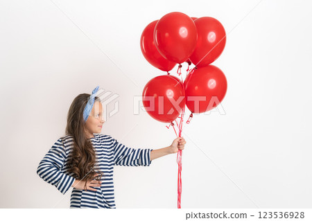Girl in striped dress holding large bunch of red balloons on outstretched hand and looking at them 123536928