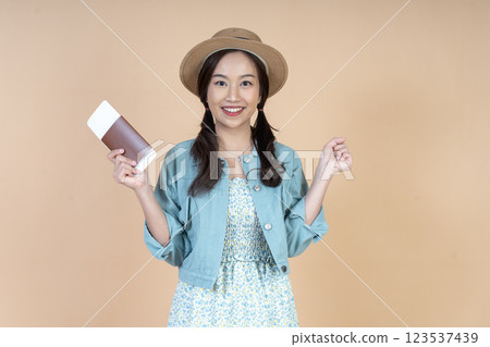 A joyful Asian woman wearing a summer dress and a straw hat holds a a passport and air ticket, posing confidently against a nude cream background in a studio with happiness. A joyful Asian woman wearing a summer dress and a straw hat holds a a passport and air ticket, posing confidently against a nude cream background in a studio with happiness. 123537439