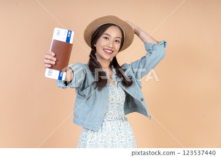A joyful Asian woman wearing a summer dress and a straw hat holds a a passport and air ticket, posing confidently against a nude cream background in a studio with happiness. 123537440