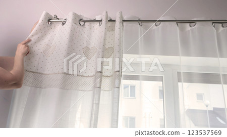 Woman hangs white curtains with metal rings made of natural material with embroidered hearts in kitchen of house. Cleaning and cleanliness. Interior and textiles.  123537569