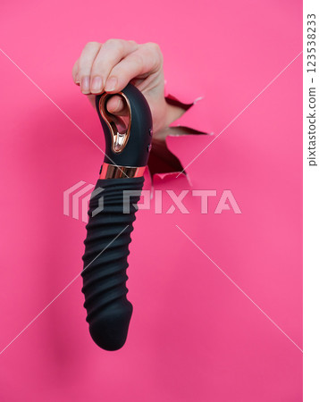 Female hand sticking out of pink paper background with black textured vibrator. Vertical photo. Female hand sticking out of pink paper background with black textured vibrator. Vertical photo. 123538233
