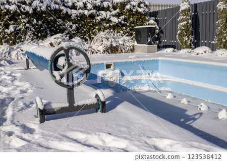 A snow-covered pool cover roller standing next to a frozen swimming pool in a winter backyard, surrounded by frost-covered trees and outdoor equipment. A snow-covered pool cover roller standing next to a frozen swimming pool in a winter backyard, surrounded by frost-covered trees and outdoor equipment. 123538412