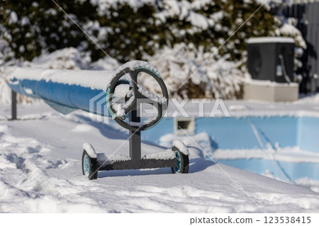 Snow-covered pool cover roller with a frozen outdoor swimming pool in the background, surrounded by a winter landscape. Snow-covered pool cover roller with a frozen outdoor swimming pool in the background, surrounded by a winter landscape. 123538415