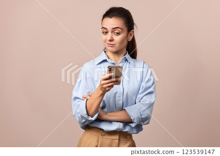 skeptical woman in blue shirt holding mobile phone looking at smartphone on beige background. 123539147