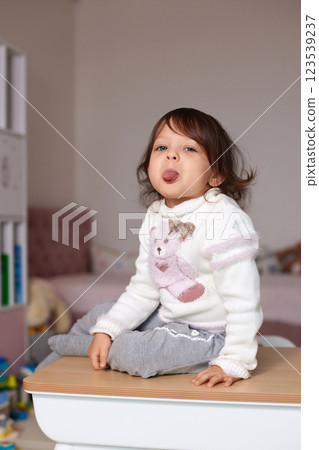 cute little child girl having fun and showing tongue while sitting on table at home cute little child girl having fun and showing tongue while sitting on table at home 123539237