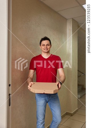 smiling delivery man in red t-shirt giving food order, holding pizza box standing at doorway smiling delivery man in red t-shirt giving food order, holding pizza box standing at doorway 123539285