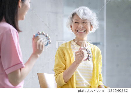 微笑的老年婦女拿著鈴鐺和護理人員音樂練習音樂治療活動圖像 微笑的老年婦女拿著鈴鐺和護理人員音樂練習音樂治療活動圖像 123540282