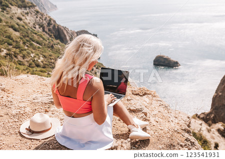 A woman is sitting on a rocky hillside with a laptop open in front of her. She is wearing a pink top and a straw hat. Concept of relaxation and leisure. 123541931