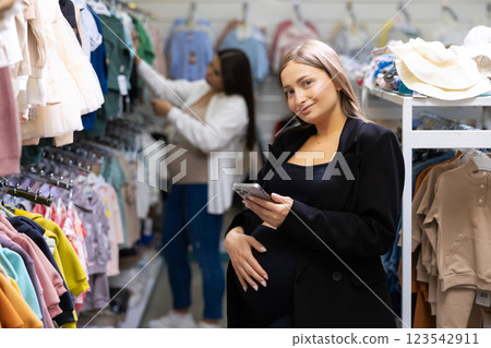 Pregnant woman choosing clothes for newborn at clothing store. Mother doing shopping in baby shop and holding mobile phone in hands. Baby clothes sale and pregnancy concept. 123542911