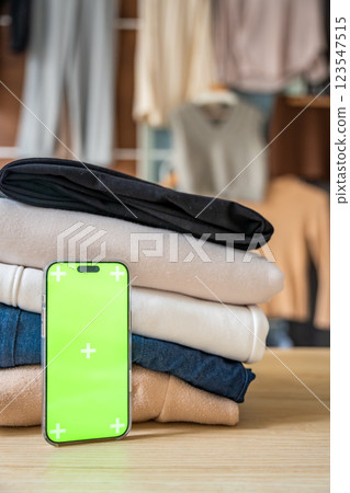 Stack of clothing prepared for resale with a smartphone displaying green screen for mockup use. Mobile apps simplify second-hand fashion, sustainable shopping, and conscious clothing redistribution 123547515