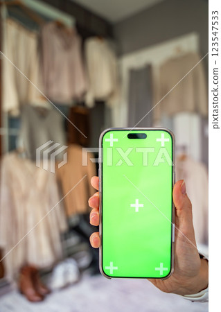 A close-up of a hand holding a smartphone with a green screen for mockup use, with blurred clothing in the background. Mobile apps and digital tools enhance online resale and second-hand fashion 123547533