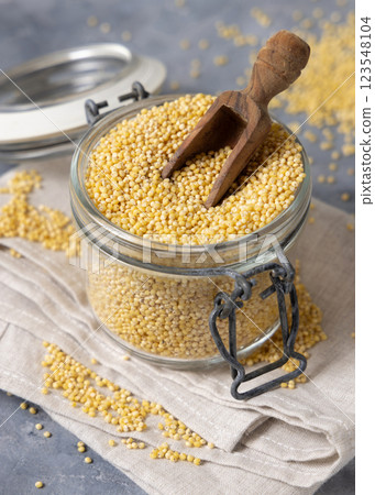 Glass jar of raw uncooked yellow millet grain with scoop on gray table closeup. Ancient food Glass jar of raw uncooked yellow millet grain with scoop on gray table closeup. Ancient food 123548104