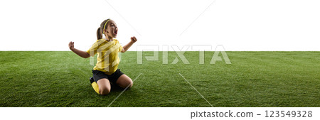 Emotional girl, soccer player in yellow jersey and black shorts sitting on grassy field, raising hands in victory pose against white background 123549328
