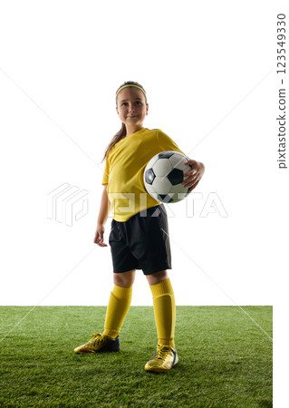 Confident girl, child in yellow jersey and black shorts proudly standing with soccer ball against white studio background. Champion 123549330