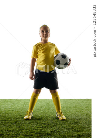 Confident girl, child in yellow jersey and black shorts proudly standing with soccer ball against white studio background. Champion 123549332