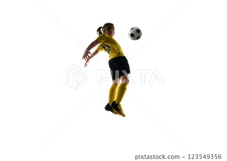Dynamic image of girl in yellow jersey and black shorts playing soccer game, kicking ball mid-air with chest against white studio background 123549356