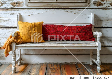 Shabby chic wooden bench with red and mustard pillows against a weathered white wooden wall, creating a cozy rustic vibe 123551875