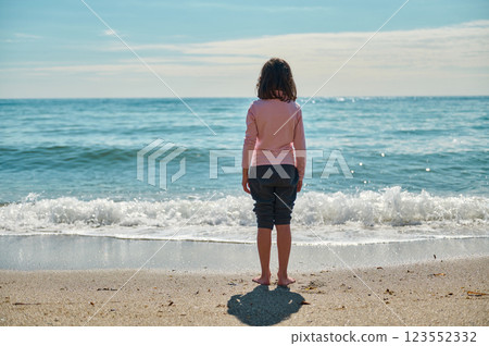 A child standing barefoot on a serene beach, appreciating the natural beauty of the calm ocean and clear summer sky. A child standing barefoot on a serene beach, appreciating the natural beauty of the calm ocean and clear summer sky. 123552332