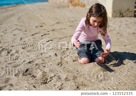 A young girl dressed in casual clothes plays alone in the sandy beach, enjoying a bright and sunny day outdoors. 123552335