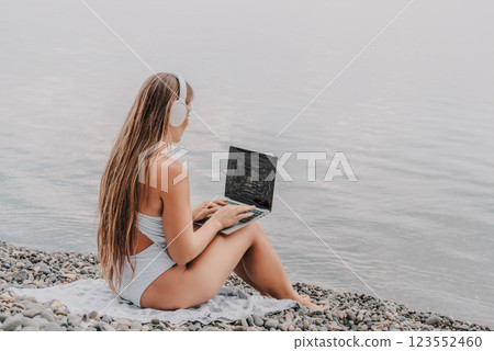 A woman is sitting on a beach with a laptop in her lap. She is wearing headphones and she is listening to music. 123552460