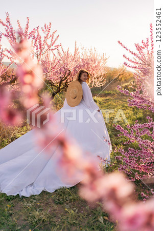 Woman blooming peach orchard. Against the backdrop of a picturesque peach orchard, a woman in a long white dress and hat enjoys a peaceful walk in the park, surrounded by the beauty of nature. 123552461