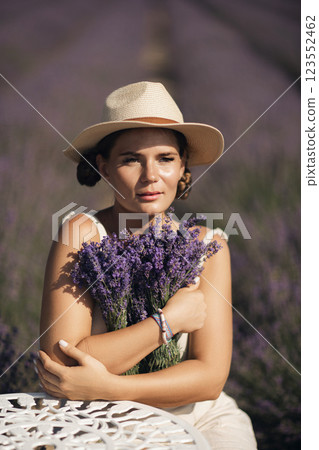 woman sitting in a field of lavender and wearing a straw hat. She is smiling and holding a bouquet of flowers. Scene is peaceful and serene, as the woman is surrounded by the beauty of nature 123552462