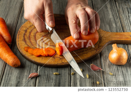 Hands skillfully slice a bright orange carrot on a wooden board, with garlic cloves and an onion nearby, showcasing a homely and engaging cooking atmosphere Hands skillfully slice a bright orange carrot on a wooden board, with garlic cloves and an onion nearby, showcasing a homely and engaging cooking atmosphere 123552730