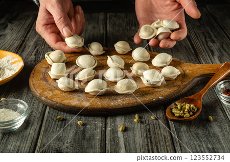Dumplings are being shaped and arranged on a wooden board, surrounded by ingredients like flour and spices, in a warm kitchen setting. The process showcases traditional cooking skills 123552734