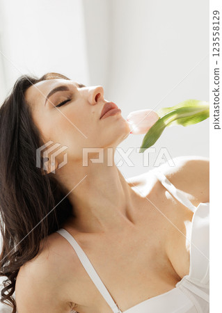 Elegant woman in white lingerie with delicate gloves holding a pink tulip near lips in soft natural light 123558129