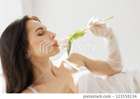 Elegant woman in white lingerie with delicate gloves holding a pink tulip near lips in soft natural light 123558134