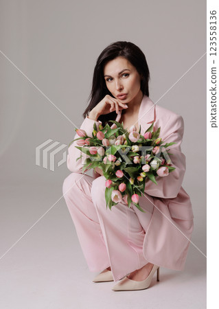 Smiling woman in a pink suit holding a bouquet of fresh tulips, posing against a neutral background in a soft, feminine, and elegant spring-inspired look 123558136