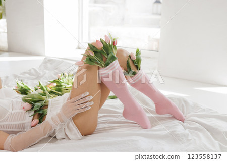 Elegant woman in lingerie lying on a bed with fresh tulips, wearing sheer gloves and pink socks, in a bright, minimalistic interior 123558137