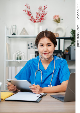 A young female nurse in blue scrubs using a digital tablet in a modern medical office setting, with a stethoscope and laptop on the desk. A young female nurse in blue scrubs using a digital tablet in a modern medical office setting, with a stethoscope and laptop on the desk. 123558988
