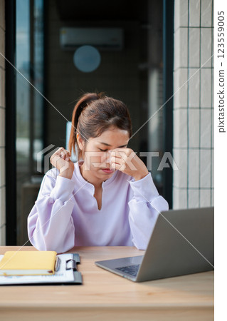 A woman in a white blouse appears stressed while sitting at a desk with a laptop and documents in a modern office environment. A woman in a white blouse appears stressed while sitting at a desk with a laptop and documents in a modern office environment. 123559005