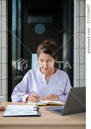 Woman sitting at a table writing in a notebook with a laptop and documents, enjoying a casual work setting. Woman sitting at a table writing in a notebook with a laptop and documents, enjoying a casual work setting. 123559007