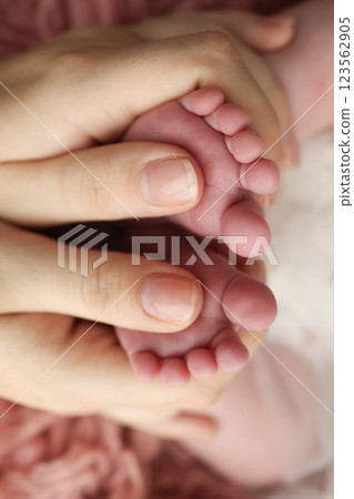 Mother holding the feet of a newborn baby on a background of pink flakatti. Feet of a newborn in the hands of parents. Macro photo of the foot, heels and toes. Openwork white dress Parents' palms. Mother holding the feet of a newborn baby on a background of pink flakatti. Feet of a newborn in the hands of parents. Macro photo of the foot, heels and toes. Openwork white dress Parents' palms. 123562905