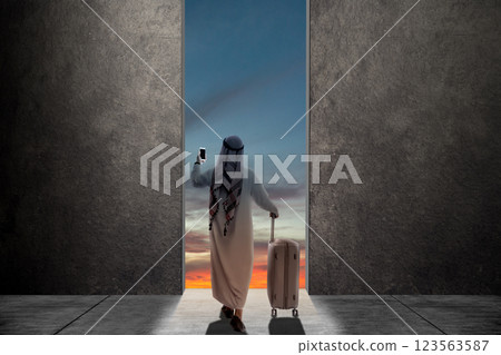 Rear view of muslim man in keffiyeh holding mobile phone while carrying suitcase. Copy space or mockup. Mudik or travel to hometown. Concept of Eid Al Fitr and Ramadan Mubarak celebration 123563587