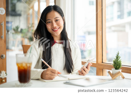 A smiling asian woman is writing in the notebook while holding the phone, sitting at the cafe table. 123564242