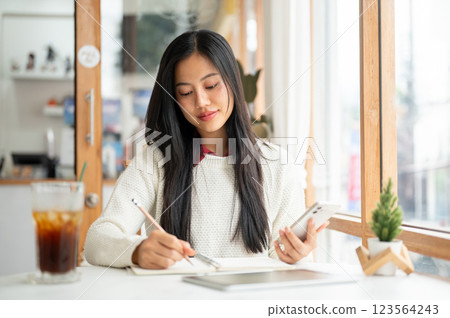An asian woman is writing in notebook while holding the phone with a tablet and a drink on the table 123564243