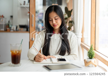 An asian woman is writing with a pencil in notebook with a smartphone and a tablet placing beside. 123564245