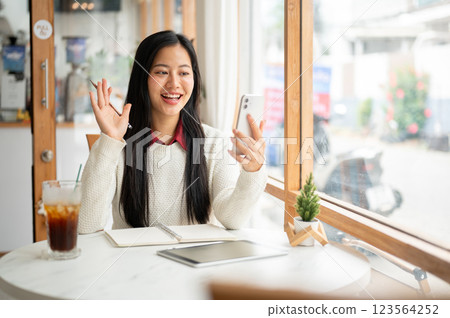 A smiling woman is waving to the phone while sitting in cafe, making a video call or live streaming. 123564252