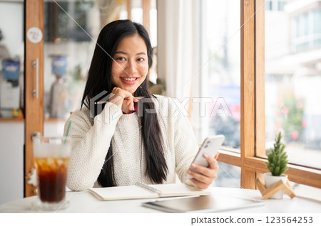 A smiling asian woman is putting one hand under the chin while holding the phone sitting in the cafe 123564253