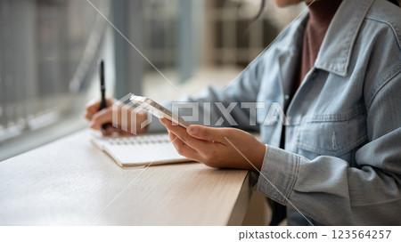 A person is looking at the smartphone while noting down or writing down in the notebook. 123564257