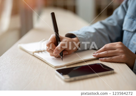 A close up of person noting down or writing down in the notebook with smartphone placing on the table A close up of person noting down or writing down in the notebook with smartphone placing on the table 123564272
