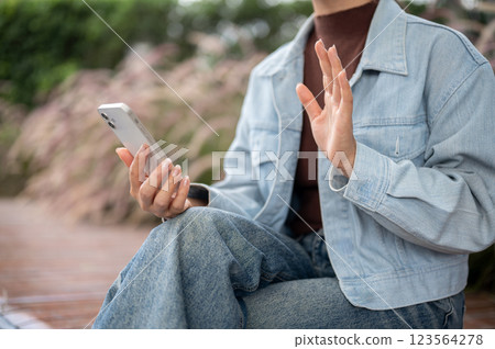 Person in denim jacket is waving to the phone while sit on bench. Video call or recording themself Person in denim jacket is waving to the phone while sit on bench. Video call or recording themself 123564278