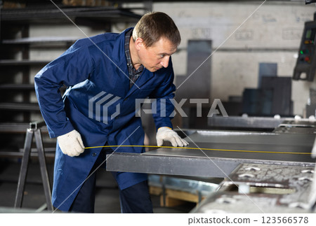 Male worker at metallurgical plant measures an iron sheet using tape measure and marks cutting line 123566578