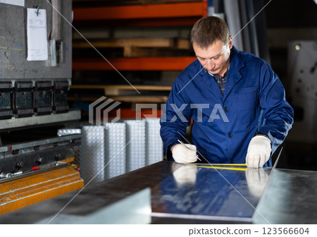 Male worker at metallurgical plant measures an iron sheet using tape measure and marks cutting line Male worker at metallurgical plant measures an iron sheet using tape measure and marks cutting line 123566604