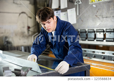 Guy working at metallurgical plant measures sheet of iron using tape measure and marks cutting line Guy working at metallurgical plant measures sheet of iron using tape measure and marks cutting line 123566634